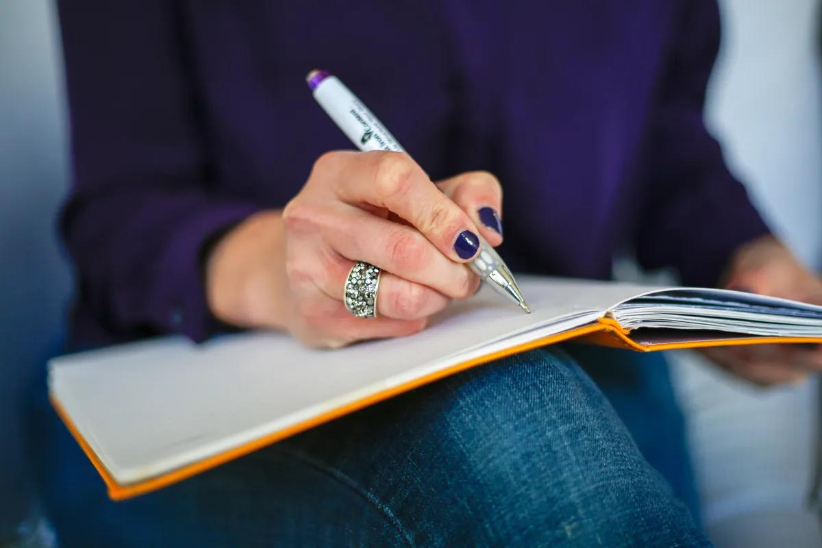 Woman writing on a notebook with a pen at a table, similar to using the included Pilot FriXion pen