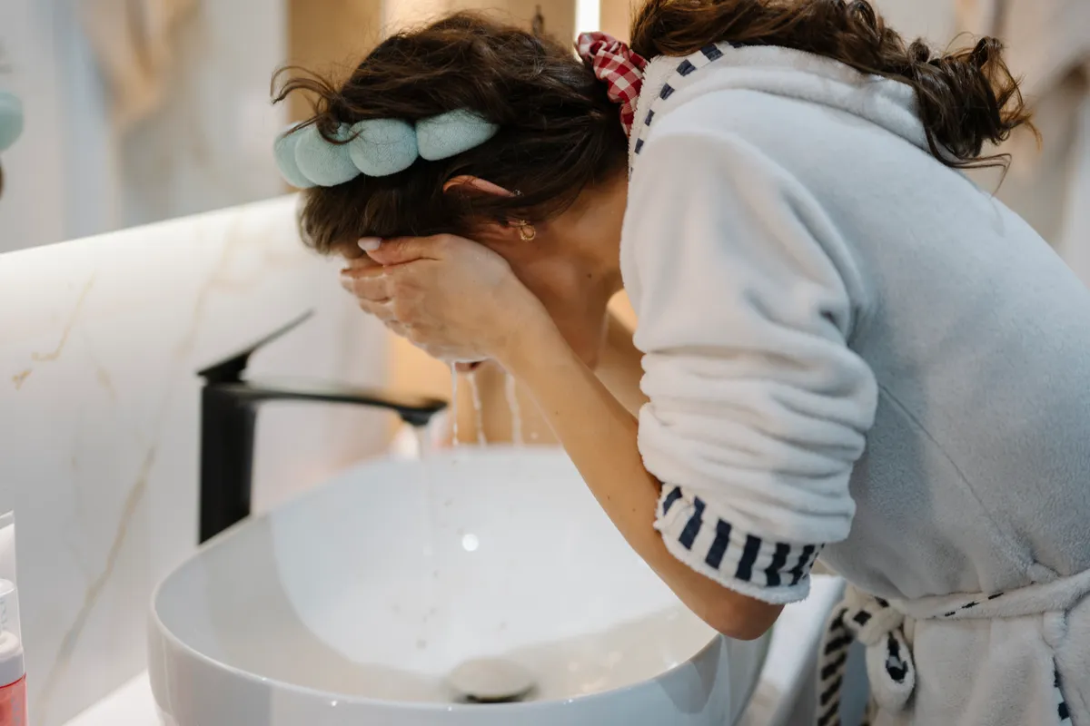 A woman splashing water on her face at a bathroom sink as part of a skincare routine